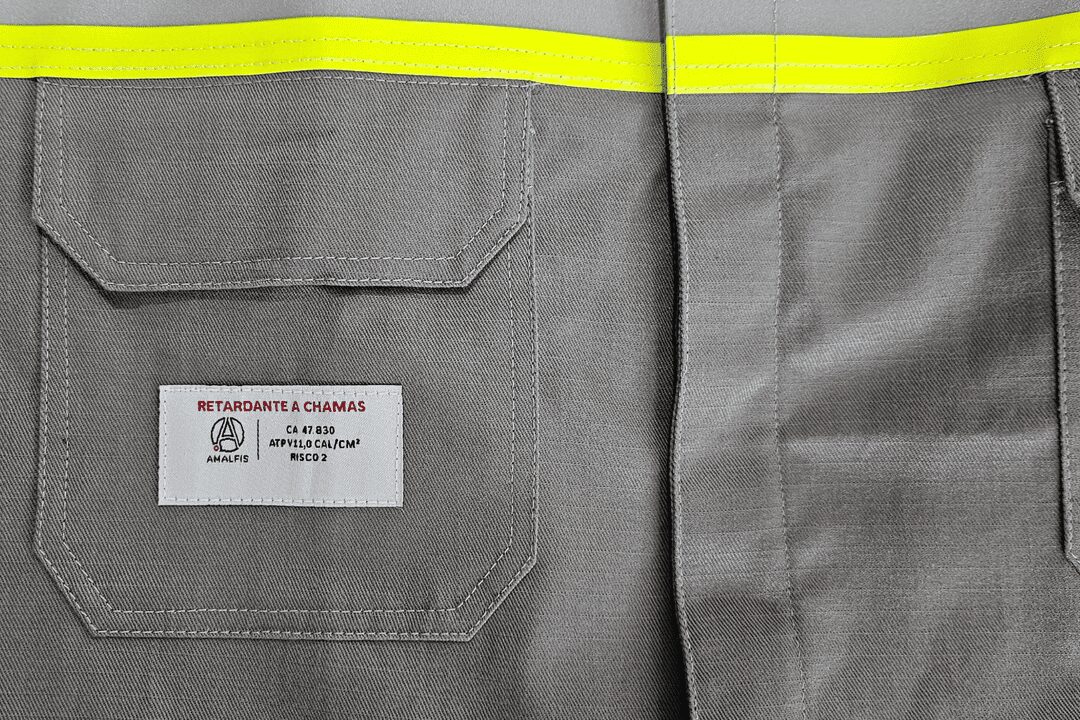 Detalhe de uniforme cinza com faixa refletiva amarela e etiqueta visível indicando o CA 47.830, classificado como retardante a chamas.