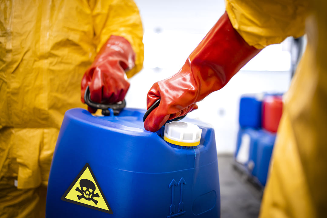 Profissionais com roupas de proteção química amarela e luvas vermelhas manuseando um galão azul com símbolo de substância tóxica.