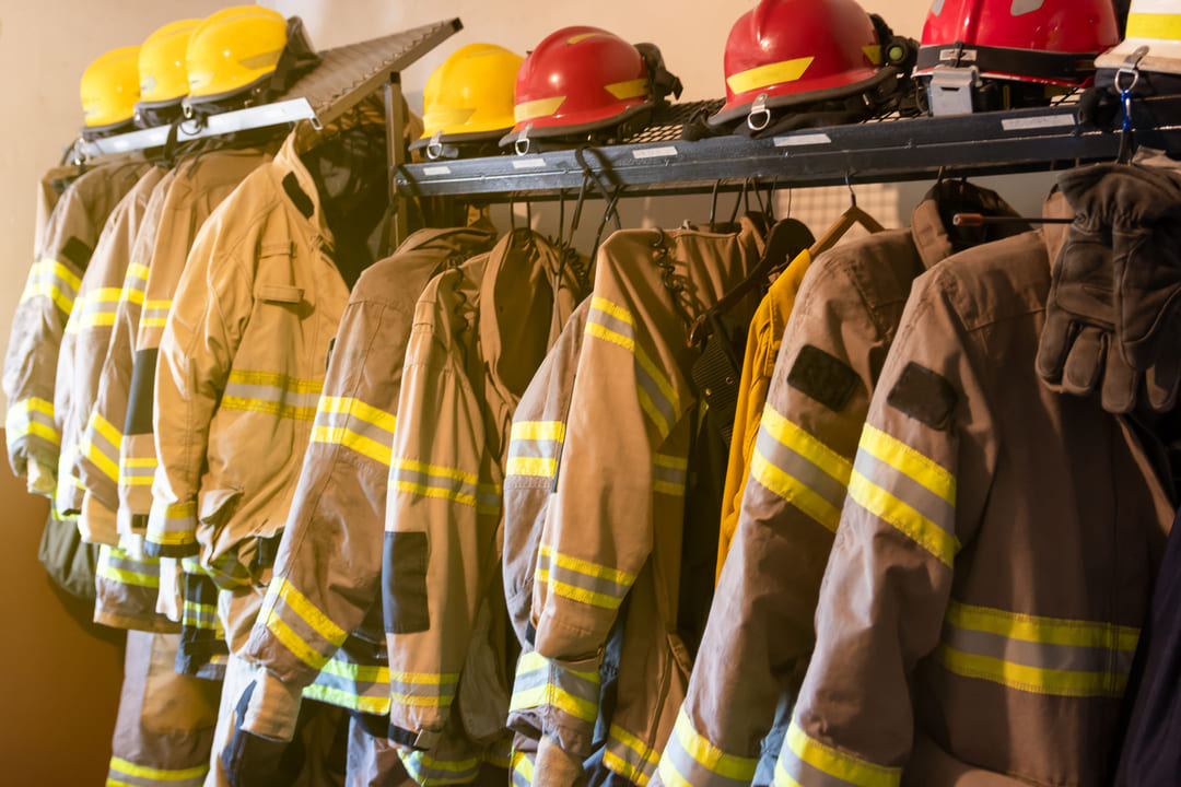 A imagem mostra um conjunto de equipamentos de proteção individual (EPIs) de bombeiros pendurados em um suporte metálico. As roupas são jaquetas resistentes de combate a incêndio, com faixas refletivas amarelas e prateadas nos braços e torso. Acima das jaquetas, há capacetes de segurança — alguns amarelos e outros vermelhos — posicionados sobre a prateleira superior do suporte. À direita, um par de luvas grossas também está pendurado. A iluminação amarelada do ambiente sugere que a foto foi tirada dentro de um quartel ou vestiário de bombeiros.