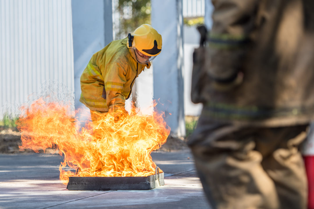 A imagem mostra uma simulação de combate a incêndio. Um bombeiro, vestindo uniforme amarelo e capacete de segurança, está curvado em frente a uma bandeja metálica com chamas intensas saindo dela. A cena ocorre em um ambiente externo, com paredes claras e piso de concreto. Em primeiro plano, à direita, parte do corpo de outro bombeiro é visível, desfocado.