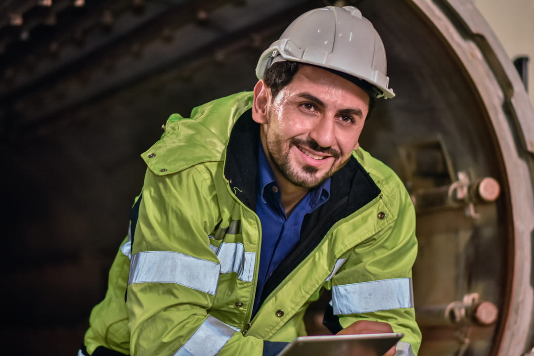 Homem sorrindo com capacete e jaqueta de alta visibilidade em ambiente industrial, ressaltando segurança NFPA 70E e NFPA 2112.