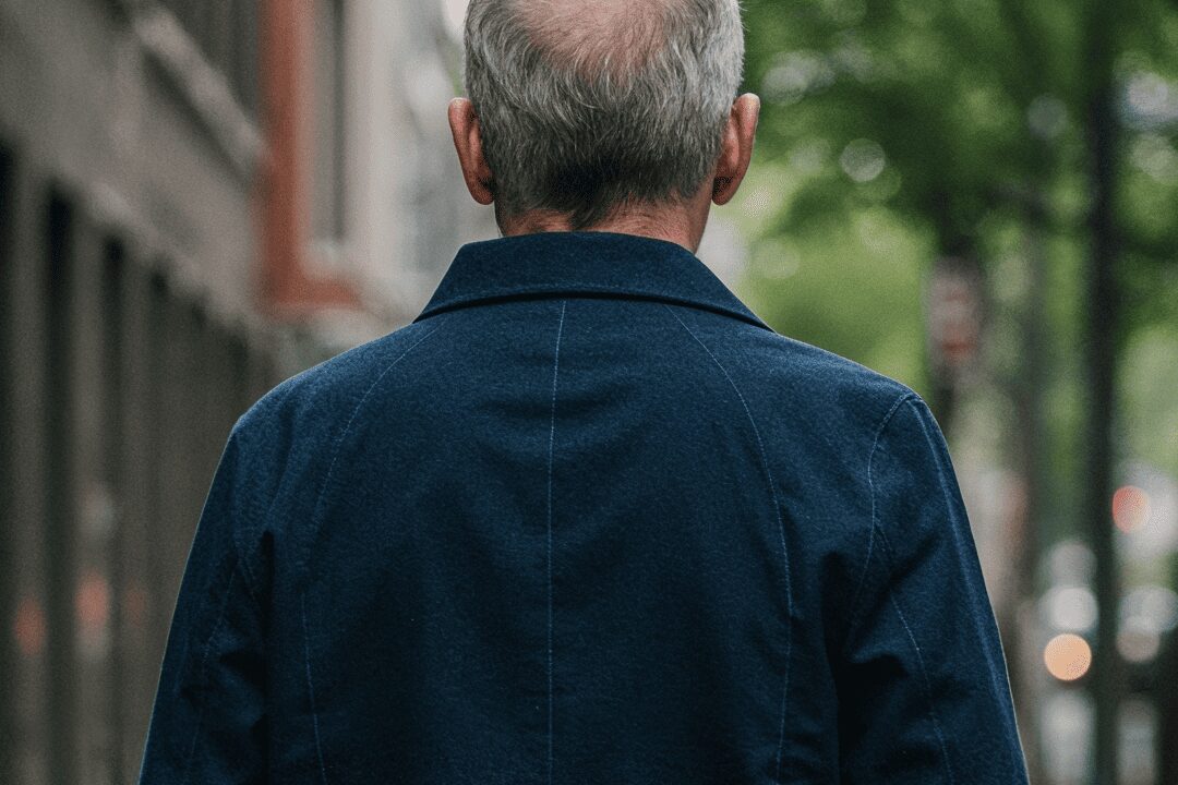 Homem visto de costas usando casaco azul feito de tecido brim, caminhando por uma rua arborizada.