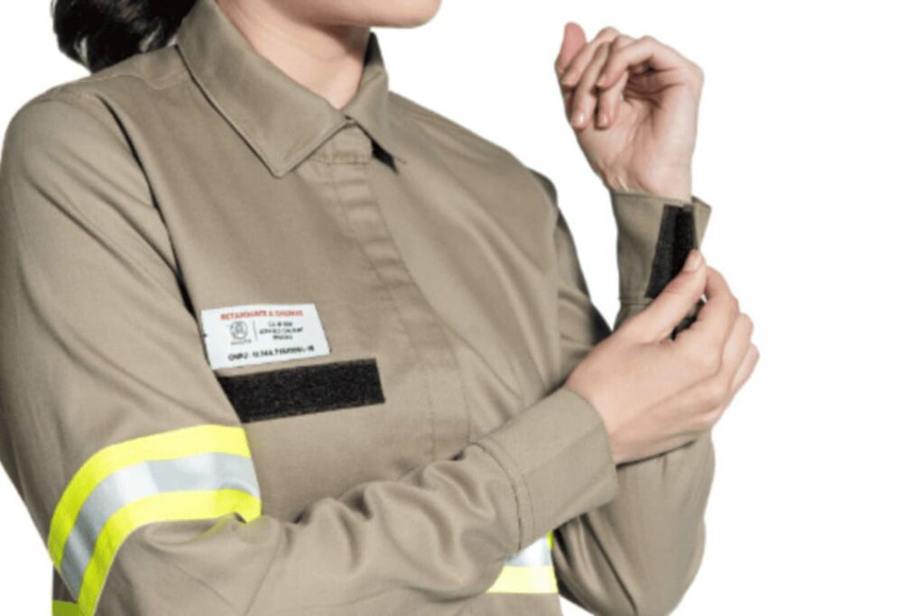 Uma pessoa em um uniforme bege, com uma faixa refletiva amarela e prata no braço, ajustando o velcro do punho da camisa. O uniforme apresenta um crachá com informações e um detalhe preto na altura do peito.