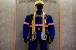 Um manequim vestido com uniforme de segurança azul com detalhes refletivos nas cores rosa e laranja, equipado com arnês de segurança amarelo. O manequim também está usando um capacete amarelo e botas pretas. O fundo consiste em uma superfície texturizada cinza com uma moldura de madeira, onde se lê parcialmente "de Segurança" no topo.