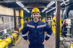 Trabalhador usando uniforme de segurança azul com faixas refletivas, capacete amarelo e protetores auriculares em ambiente industrial com tubulações e maquinário.
