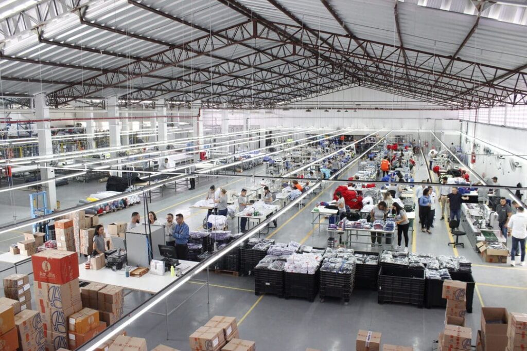 Linha de produção têxtil com costureiros trabalhando em uniformes corporativos dentro de uma ampla fábrica organizada.