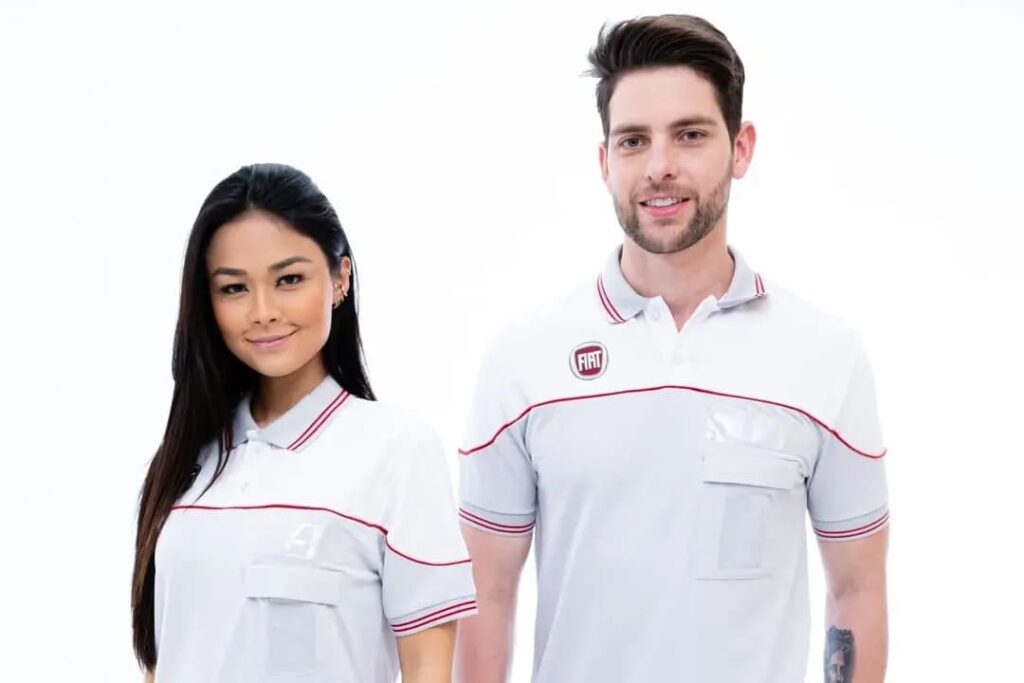 Um homem e uma mulher estão vestidos com uniformes brancos com detalhes em vermelho e o logo da Fiat no lado esquerdo do peito. Eles estão posicionados lado a lado, sorrindo, com um fundo branco neutro.