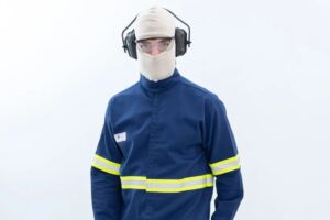 Uma pessoa vestindo um uniforme azul com detalhes de faixas refletivas amarelas nos braços e na cintura. Ela também está usando um capuz de proteção bege que cobre toda a cabeça, deixando apenas os olhos visíveis através de óculos de segurança transparentes. Além disso, está equipada com protetores auriculares pretos. O fundo é branco.