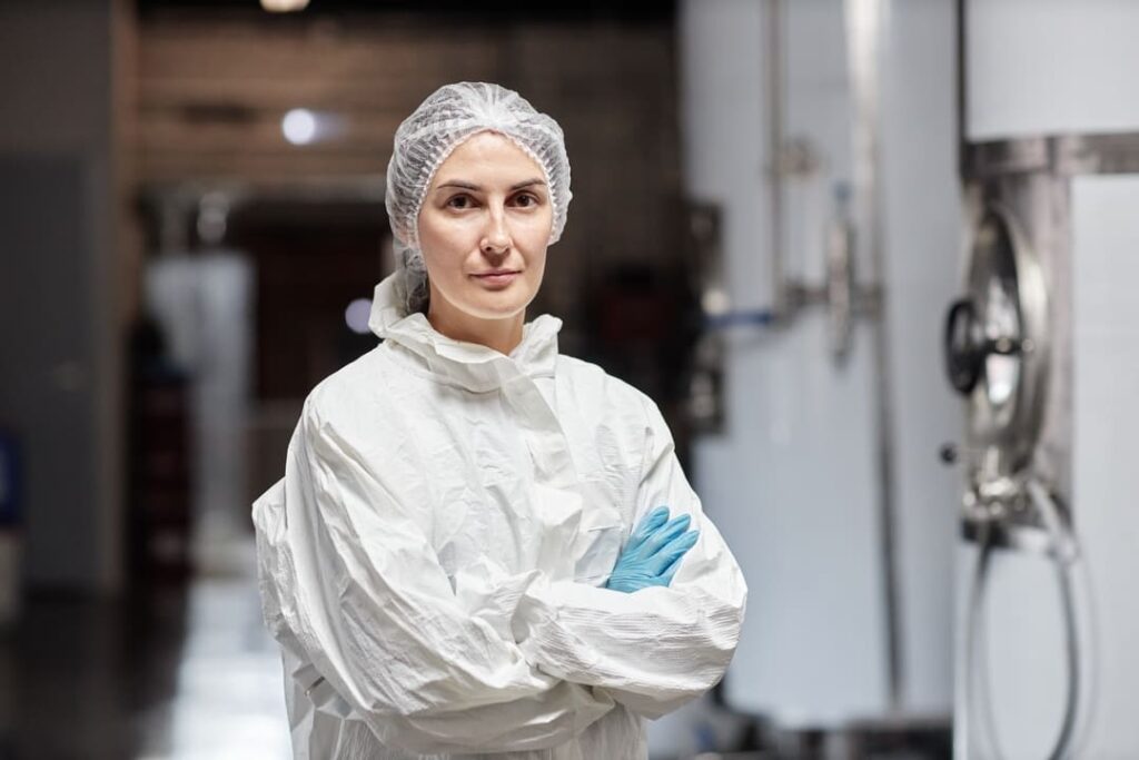 Profissional usando vestimenta de proteção química branca com touca e luvas azuis em ambiente industrial.