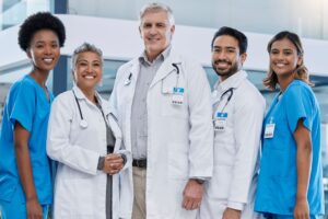 Equipe médica sorridente, composta por médicos e enfermeiros com jalecos e crachás, posando juntos em ambiente hospitalar moderno.