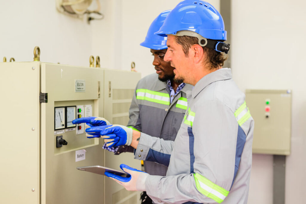 Técnicos com EPI inspecionam painel elétrico em ambiente industrial.