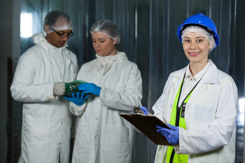 Profissionais em ambiente industrial com jalecos brancos, toucas descartáveis, luvas e capacetes, seguindo normas de vestimentas de proteção.