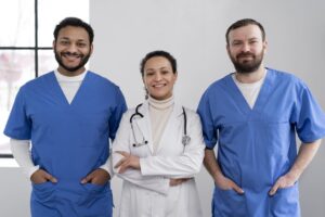 Equipe médica sorridente com dois profissionais usando scrubs azuis e uma médica ao centro com jaleco branco e estetoscópio.