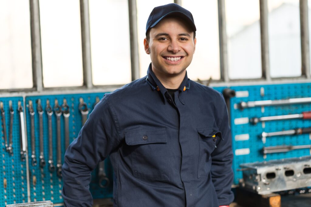 Homem sorridente vestindo uniforme azul-marinho e boné, posando em frente a um painel com ferramentas em uma oficina mecânica.